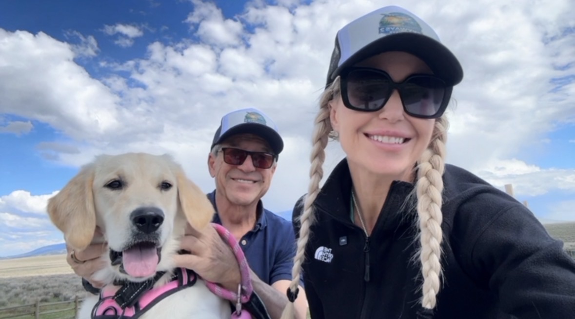 Kristie and Fred smiling with their golden retriever Goldie on a grassy plain